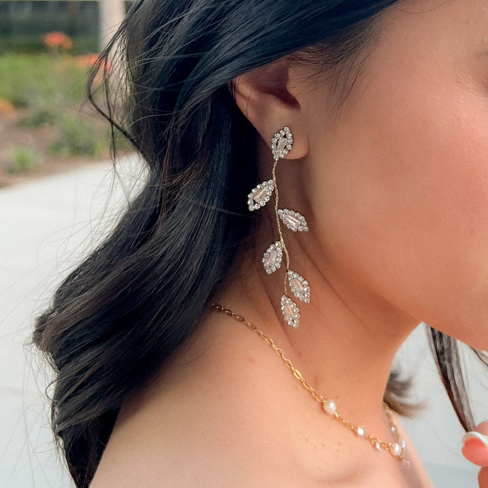 A close-up image of a woman wearing gold-tone Juniper earrings with rhinestone embellishments, featuring a cascading leaf design.