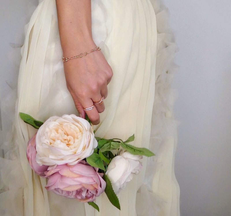 A person in a cream-colored dress holds three light pink and white peonies with green leaves. The person is wearing a delicate bracelet and rings on their left hand.