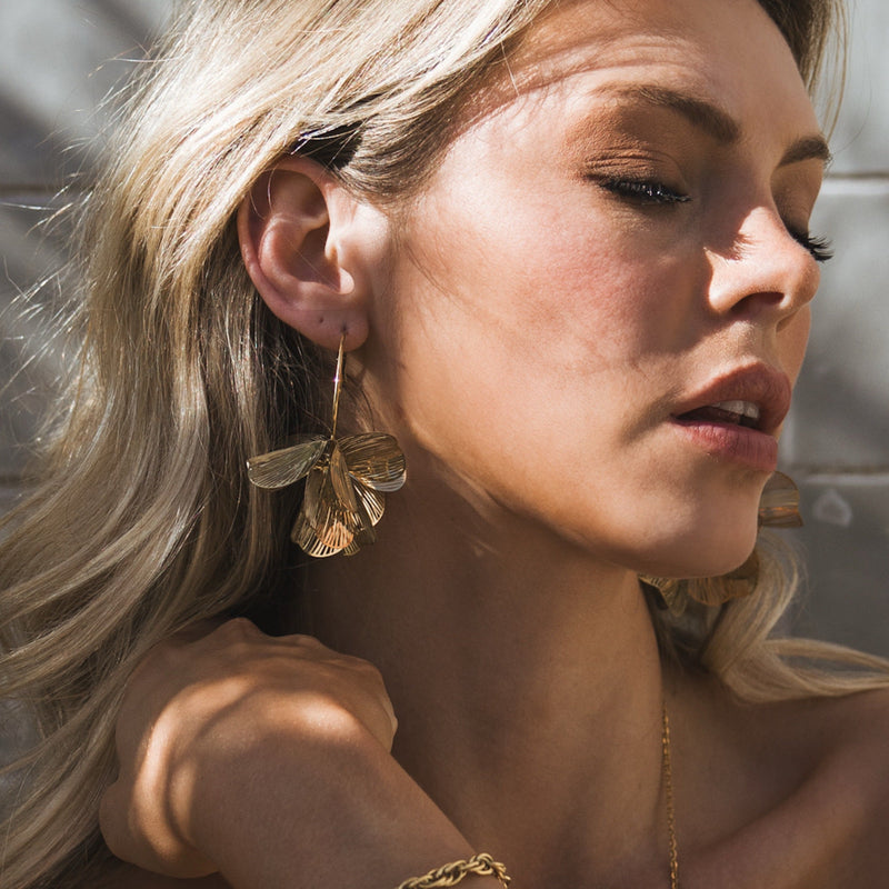 A woman with long blonde hair poses in sunlight, eyes closed, wearing large gold butterfly earrings and a delicate gold necklace. Shadows and light create texture on her face and background.