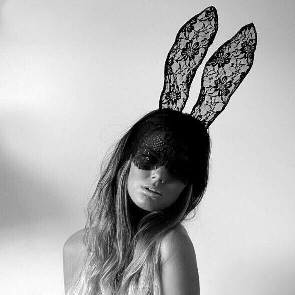 A woman with long hair wears a black lace mask and matching lace bunny ears, posing against a plain light background in a black and white photo.