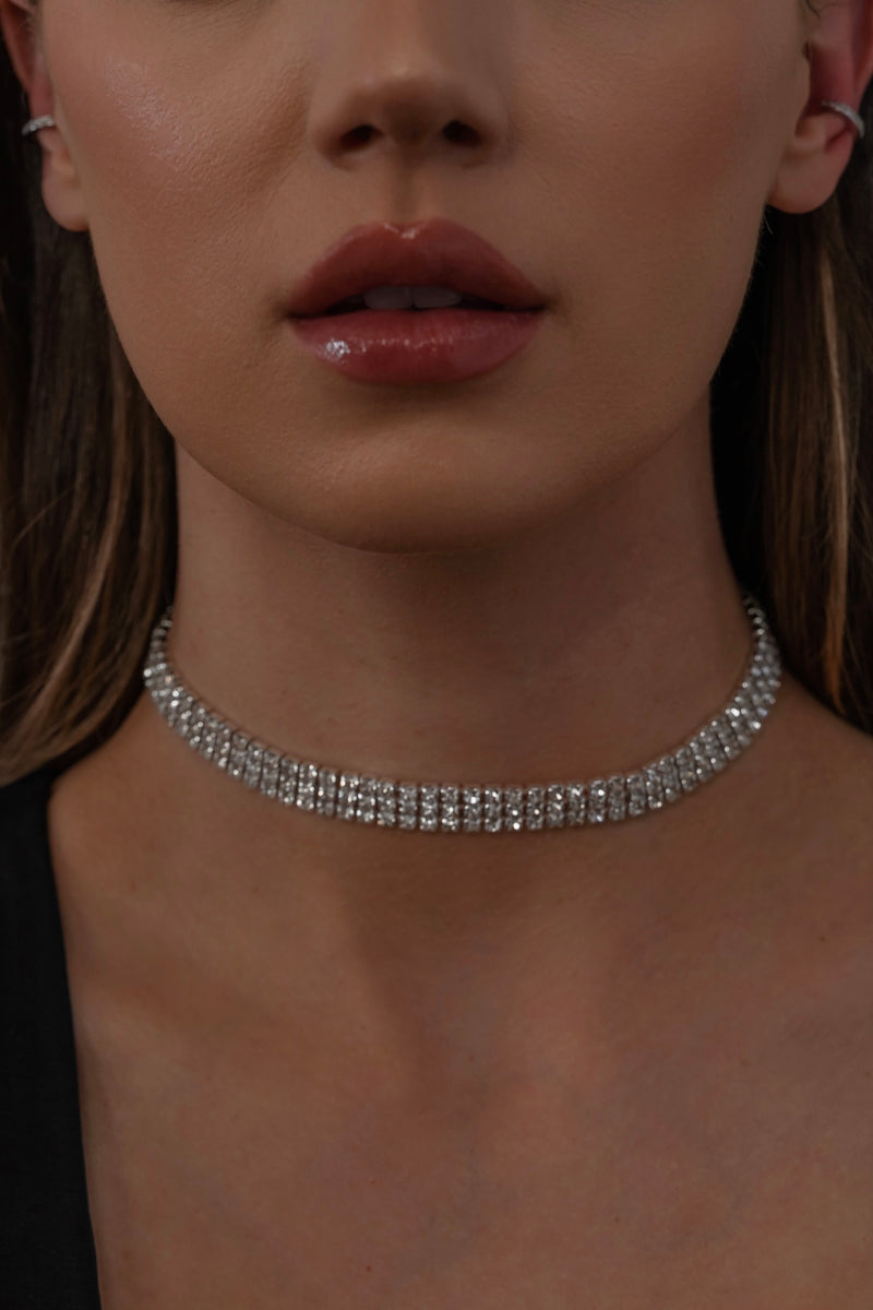 A close-up of a woman wearing a sparkling, multi-row rhinestone choker necklace and a black top. Her lips are slightly parted and her skin has a smooth, even complexion.