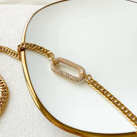 The Songbird Collections Icon Link Necklace, featuring a gold chain and rectangular, crystal-encrusted pendant, is showcased on a white plate with a gold rim to elegantly highlight its shine and texture.