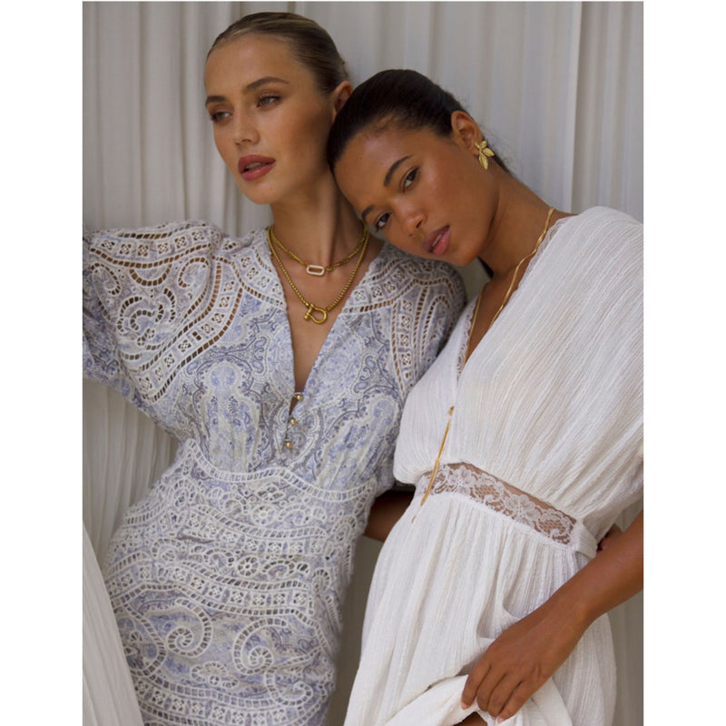 Two women stand against a soft, light-colored curtain backdrop in elegant white dresses with lace details. One leans gently on the others shoulder, both wearing gold jewelry, including The Songbird Collections Icon Link Necklace.