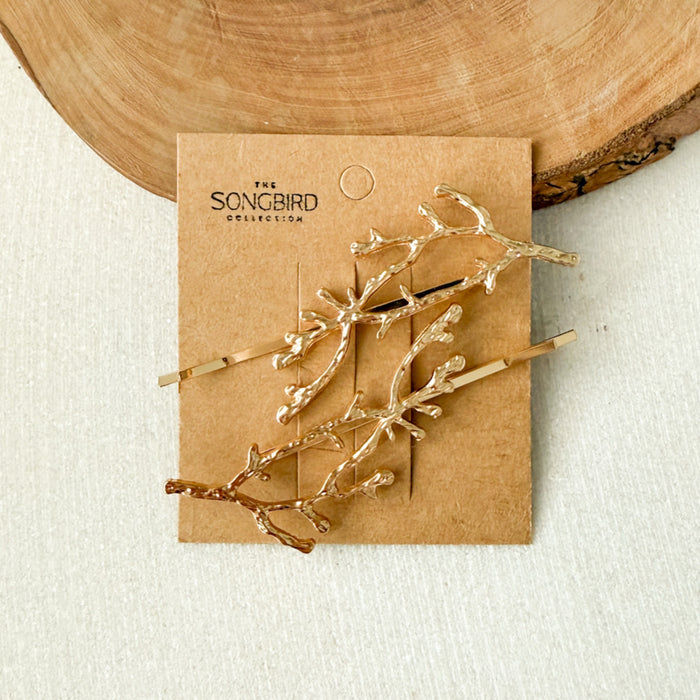 Three gold branch-shaped hair pins from The Songbird Collection, displayed on a brown kraft paper backing card against a light textured background with a wood slice prop.  Arbors Hair Pin Set - The Songbird Collection
