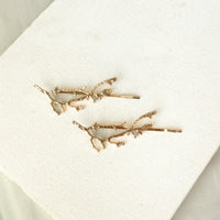 A pair of gold branch-shaped hair pins displayed on a textured white fabric background.  Arbors Hair Pin Set - The Songbird Collection