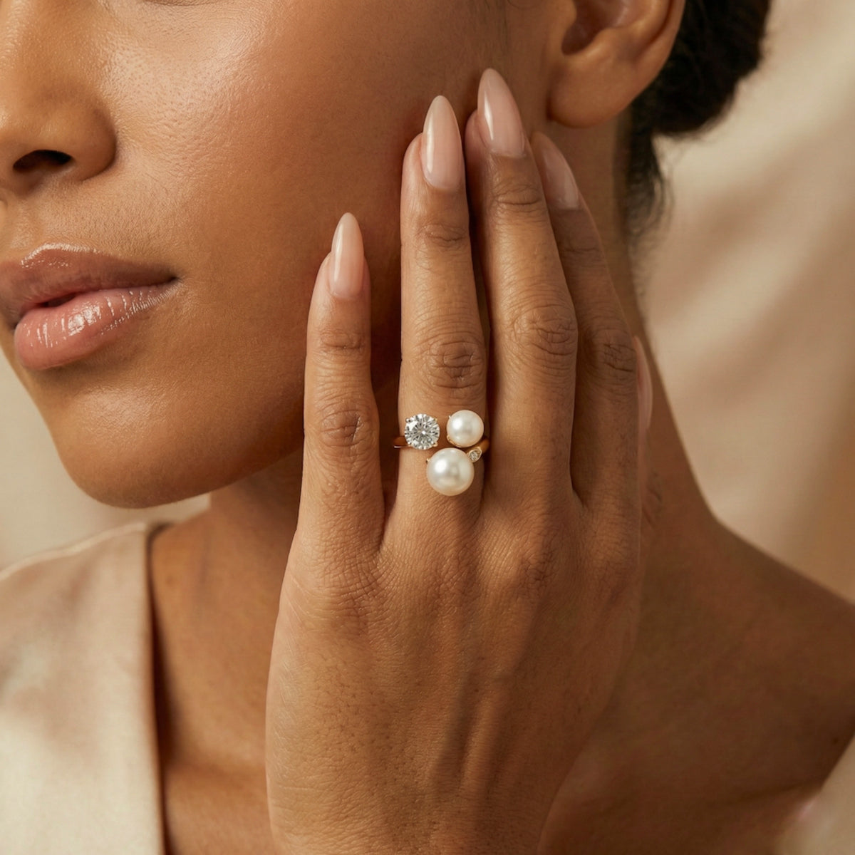 Close-up of a woman's hand wearing a ring with pearls and a diamond, touching her face.