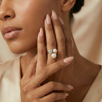 Close-up of a woman's hand wearing a ring with pearls and a diamond, against a neutral background.