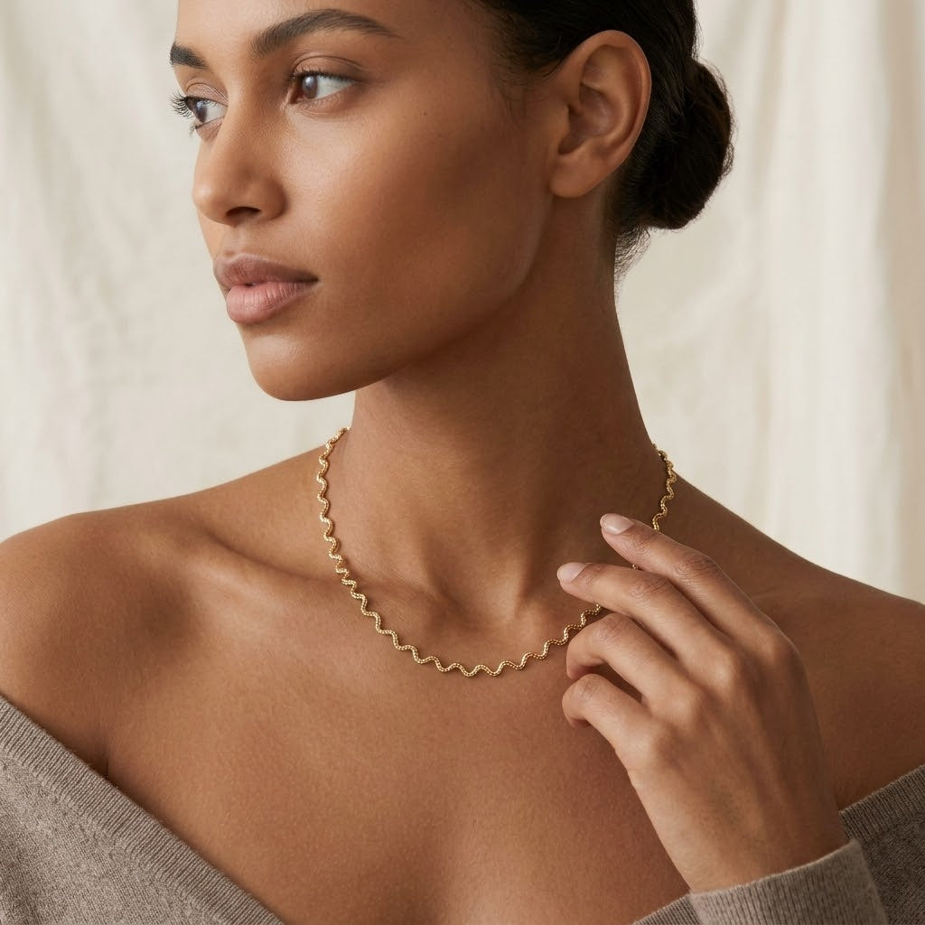 A woman wearing a delicate gold wavy chain necklace, shown in a close-up profile view against a soft, light beige background. Aura_Ripple_Chain_Necklace_-_The_Songbird_Collection