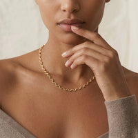 A close-up of a model wearing a delicate gold wavy chain necklace, with her hand resting near her chin against a soft, light beige background.