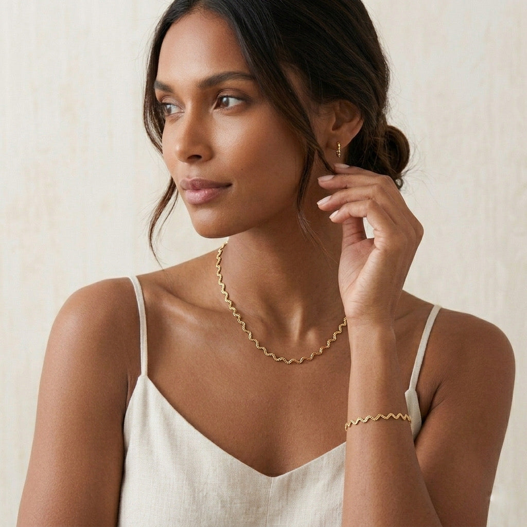 Woman wearing gold jewelry including a necklace and bracelet against a neutral background - stainless steel silver wavy chain necklace a marble surface - Aura_Ripple_Chain_Necklace_-_The_Songbird_Collection 