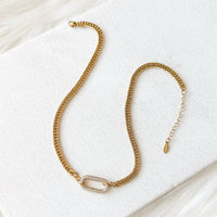 The Songbird Collections Icon Link Necklace is a gold choker with a rectangular pavé crystal centerpiece, showcased on a white textured surface against a soft, light background.