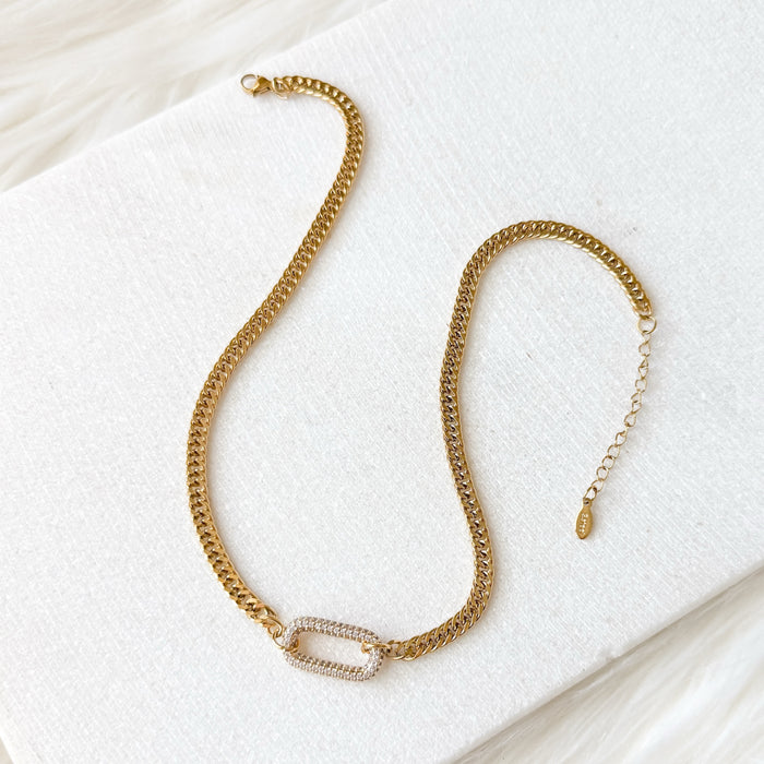 The Songbird Collections Icon Link Necklace is a gold choker with a rectangular pavé crystal centerpiece, showcased on a white textured surface against a soft, light background.
