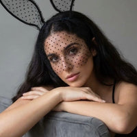 A woman with long dark hair wears a black polka-dot veil over her face and black bunny ears on a headband, leaning her arms on a gray armchair against a plain gray background. Black_Lace_Polka_Dot_Bunny_Ear_Headband_-_The_Songbird_Collection