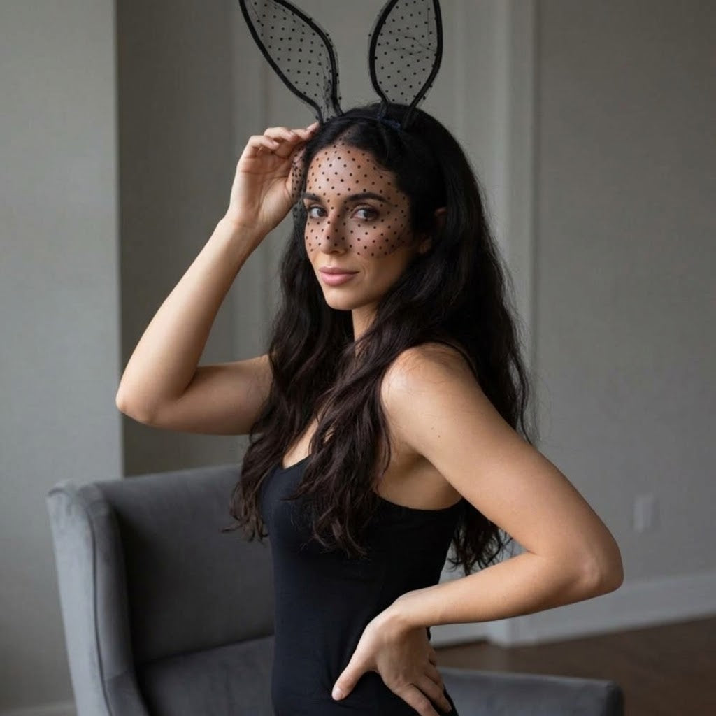 A woman wearing a black bunny costume accessory set, including black polka-dot bunny ears on a headband and matching black polka-dot face veil, posing in a black bodysuit in a gray armchair. Black_Lace_Polka_Dot_Bunny_Ear_Headband_-_The_Songbird_Collection