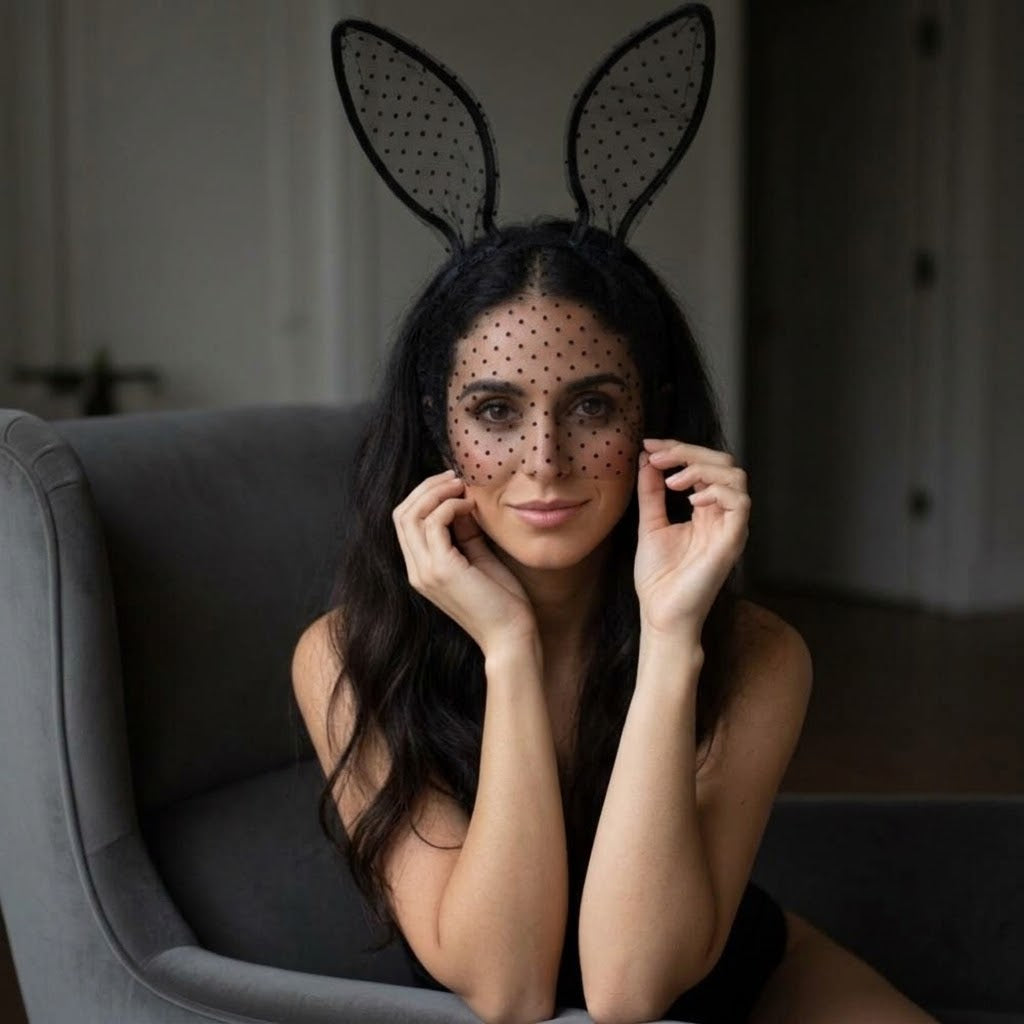 A woman wearing a black bunny headband with long ears and a matching black polka-dot veil over her eyes, sitting in a gray armchair in a dimly lit room. Black_Lace_Polka_Dot_Bunny_Ear_Headband_-_The_Songbird_Collection