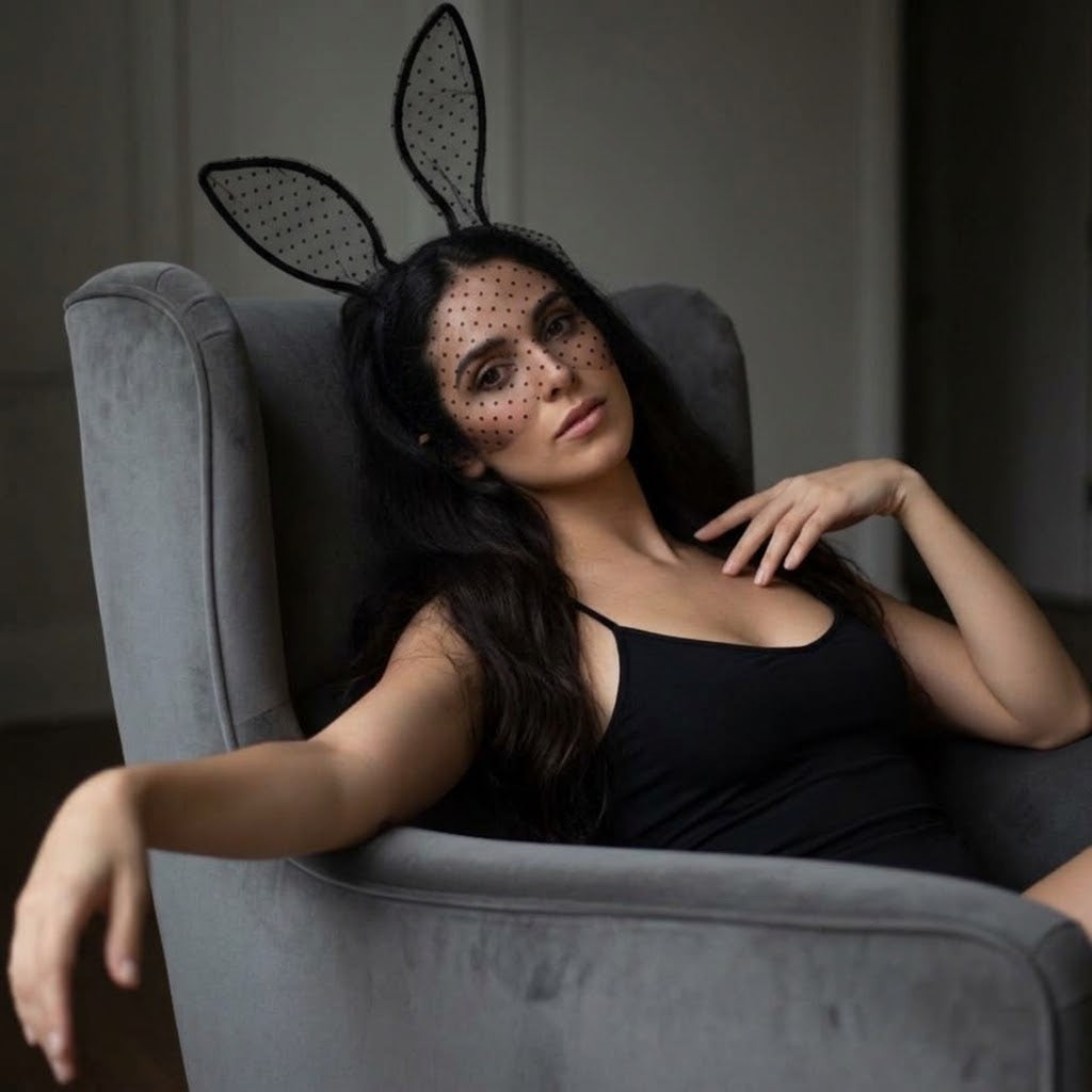 A woman reclining in a gray armchair wearing a black lace bunny headband with black polka-dot mesh and a black camisole.  Black_Lace_Polka_Dot_Bunny_Ear_Headband_-_The_Songbird_Collection