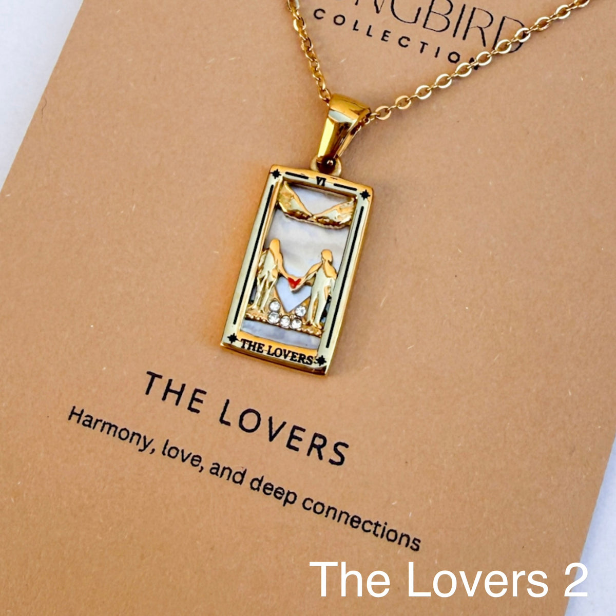 Gold pendant necklace with 'The Lovers' design on a brown card, featuring the brand 'The Songbird Collection'.