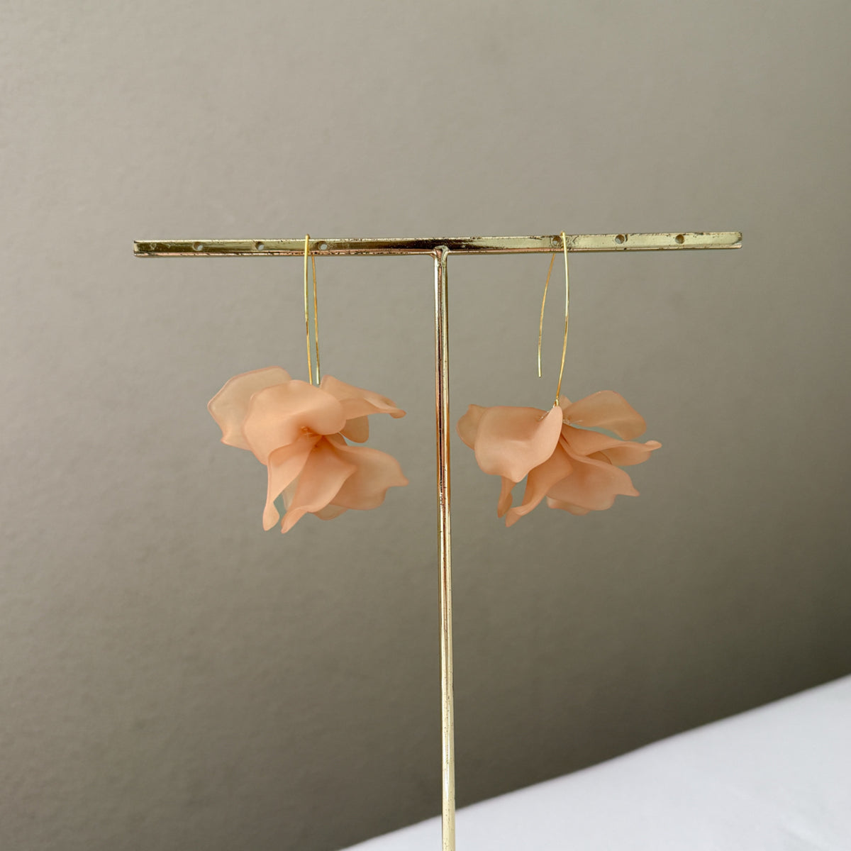 Pair of peach-colored flower earrings on a gold earring stand against a beige background.  Cyclamen Petal Drop Earrings - Chai Tea Latte - The Songbird Collection