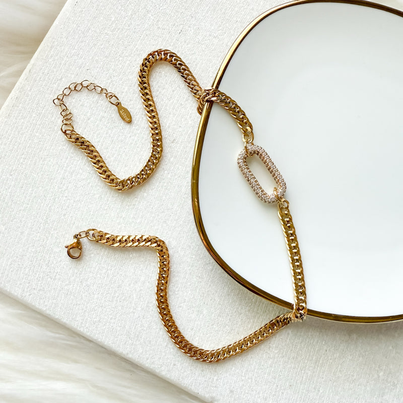 The Songbird Collection’s Icon Link Necklace, featuring a sparkling oval pavé crystal pendant, is elegantly draped over a gold-rimmed white plate on a textured white surface.
