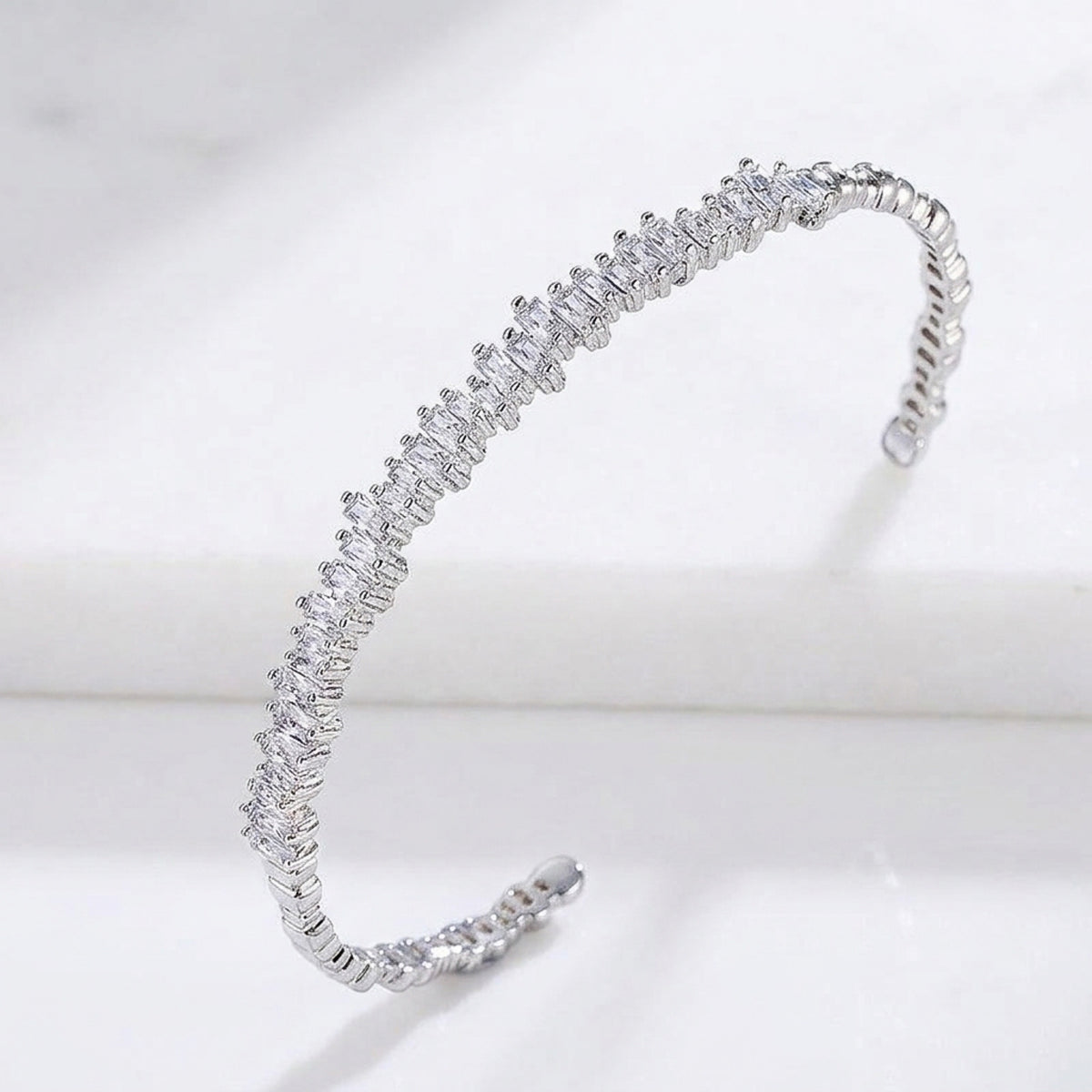 A silver-toned open cuff bracelet adorned with a continuous row of small clear marquise-cut stones, displayed on a white marble surface.  Esra Open Bangle - The Songbird Collection 