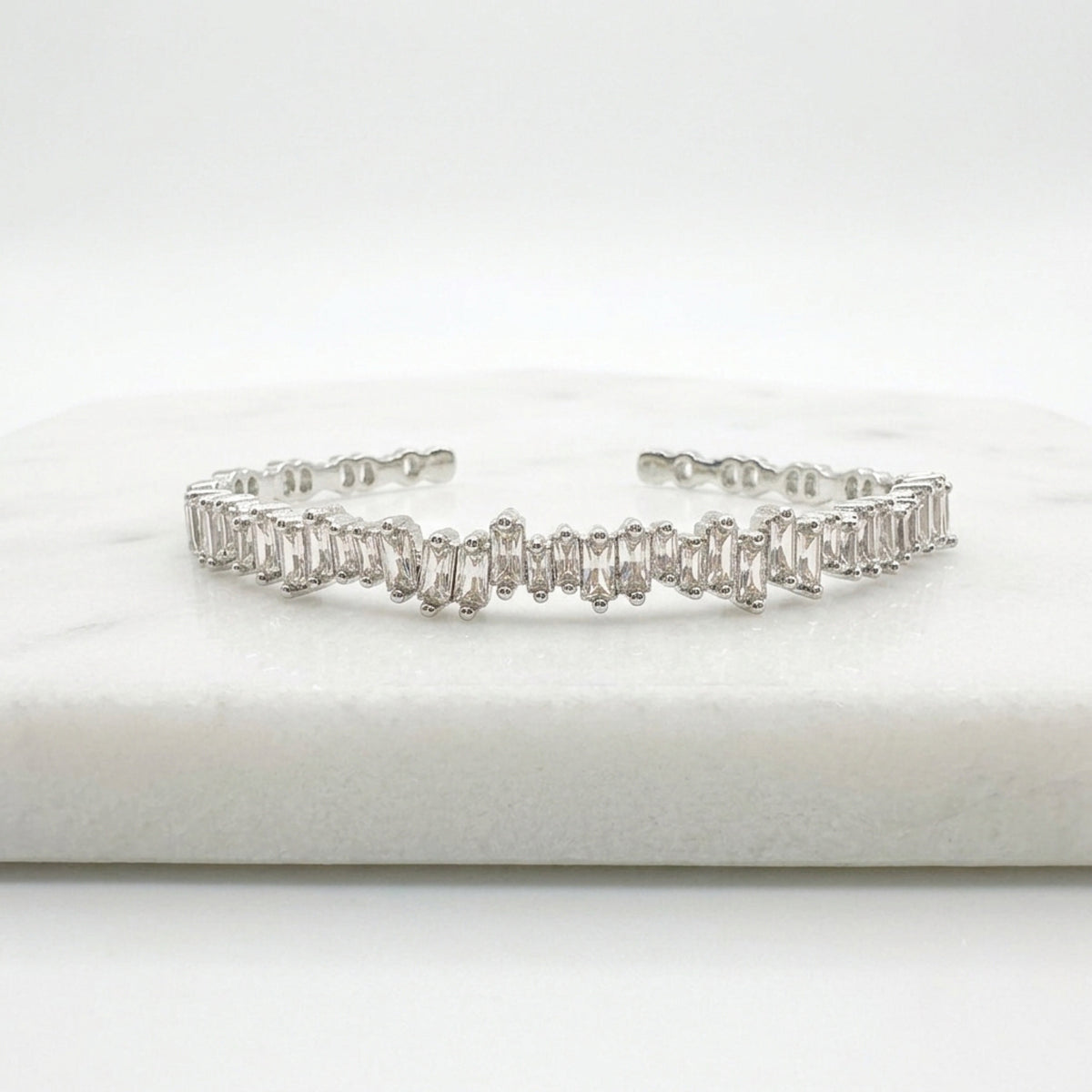 A silver-toned cuff bracelet featuring a row of baguette-cut clear stones, displayed on a white marble block against a white background. Esra Open Bangle - The Songbird Collection 