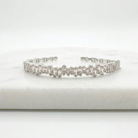 A silver-toned cuff bracelet featuring a row of baguette-cut clear stones, displayed on a white marble block against a white background. Esra Open Bangle - The Songbird Collection 