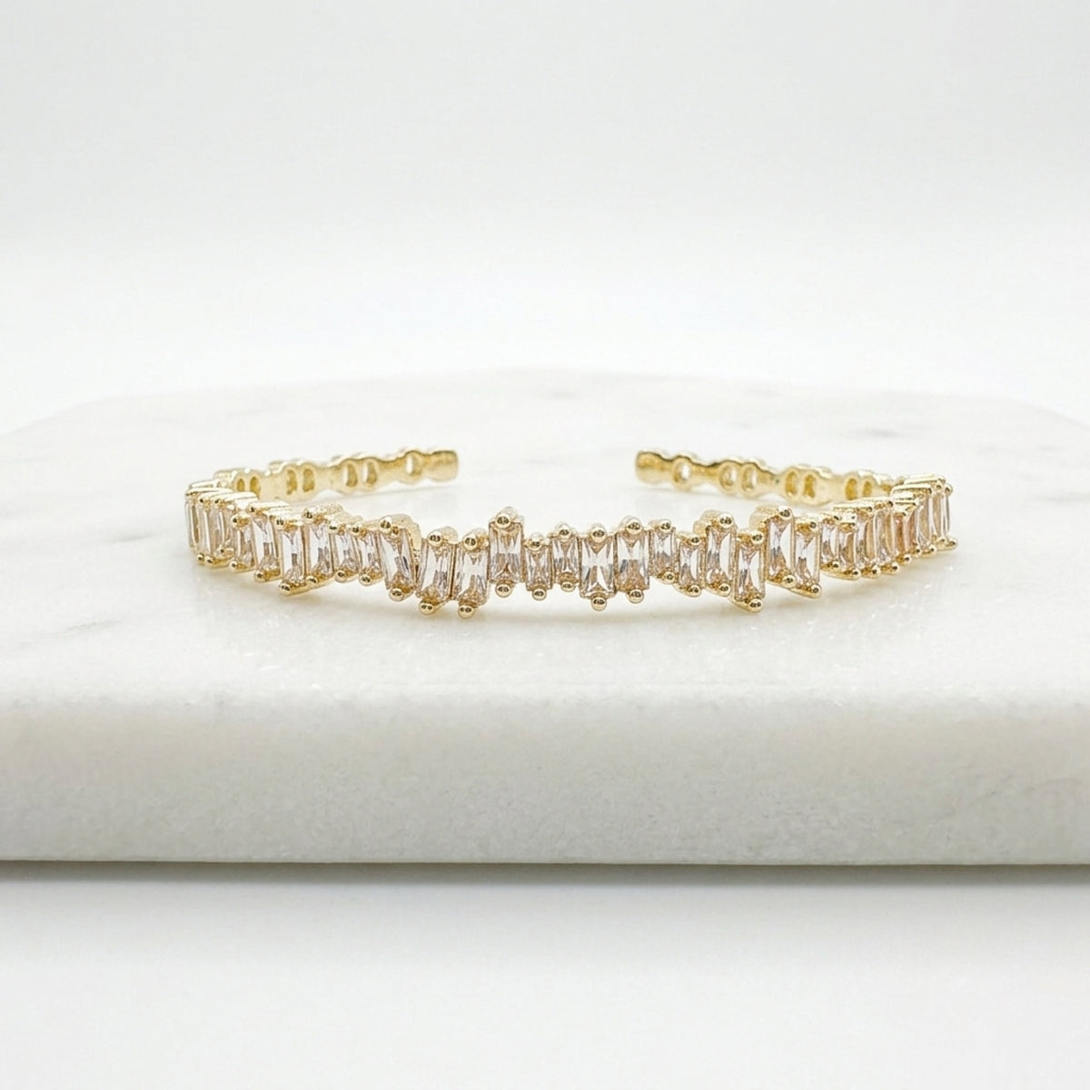 A gold-toned cuff bracelet featuring a row of clear baguette-cut stones, displayed on a white marble block against a white background.  Esra Open Bangle - The Songbird Collection 