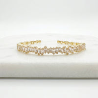 A gold-toned cuff bracelet featuring a row of clear baguette-cut stones, displayed on a white marble block against a white background.  Esra Open Bangle - The Songbird Collection 