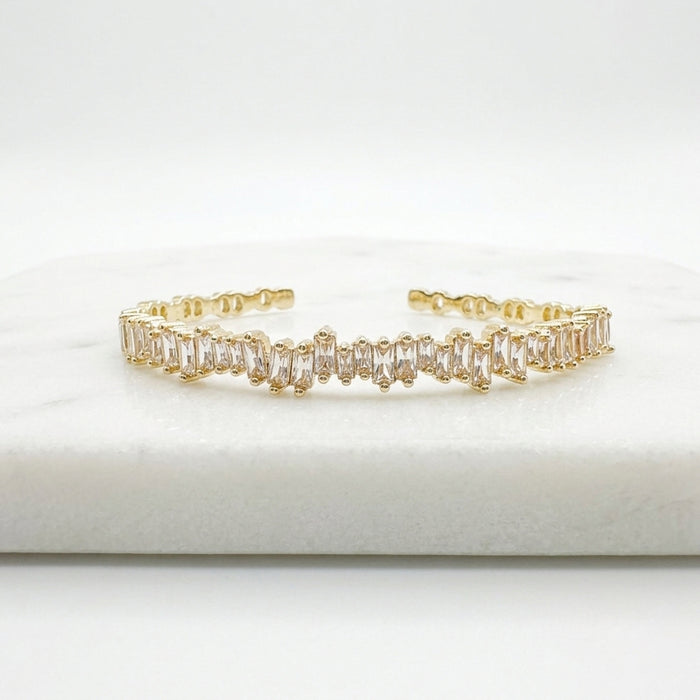 A gold-toned cuff bracelet featuring a row of clear baguette-cut stones, displayed on a white marble block against a white background.  Esra Open Bangle - The Songbird Collection 