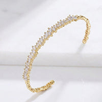 A gold open cuff bracelet featuring a curved row of clear round-cut stones, displayed on a white marble surface. Esra Open Bangle - The Songbird Collection 