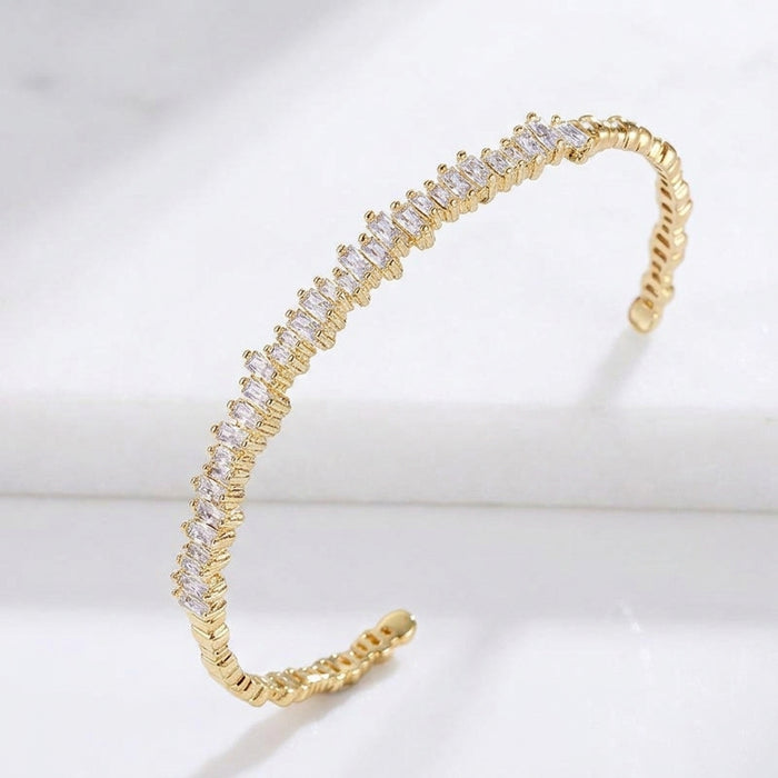 A gold open cuff bracelet featuring a curved row of clear round-cut stones, displayed on a white marble surface. Esra Open Bangle - The Songbird Collection 