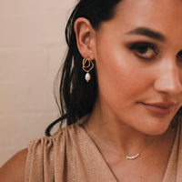A woman wearing a beige top and a minimalist style pearl earring with gold plating.  Faith Freshwater Pearl Earrings - The Songbird Collection 