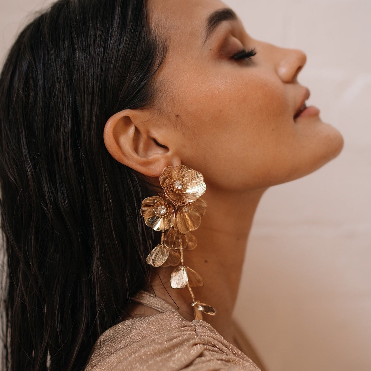 A woman wearing golden floral statement earrings with intricate details and dangling chains, posing with her eyes closed.