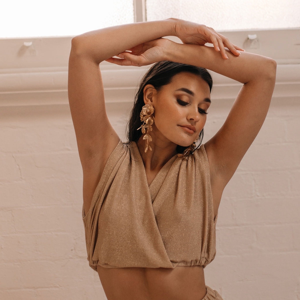 A woman wearing golden floral statement earrings and a beige top, posing with her arms raised and hands clasped above her head.