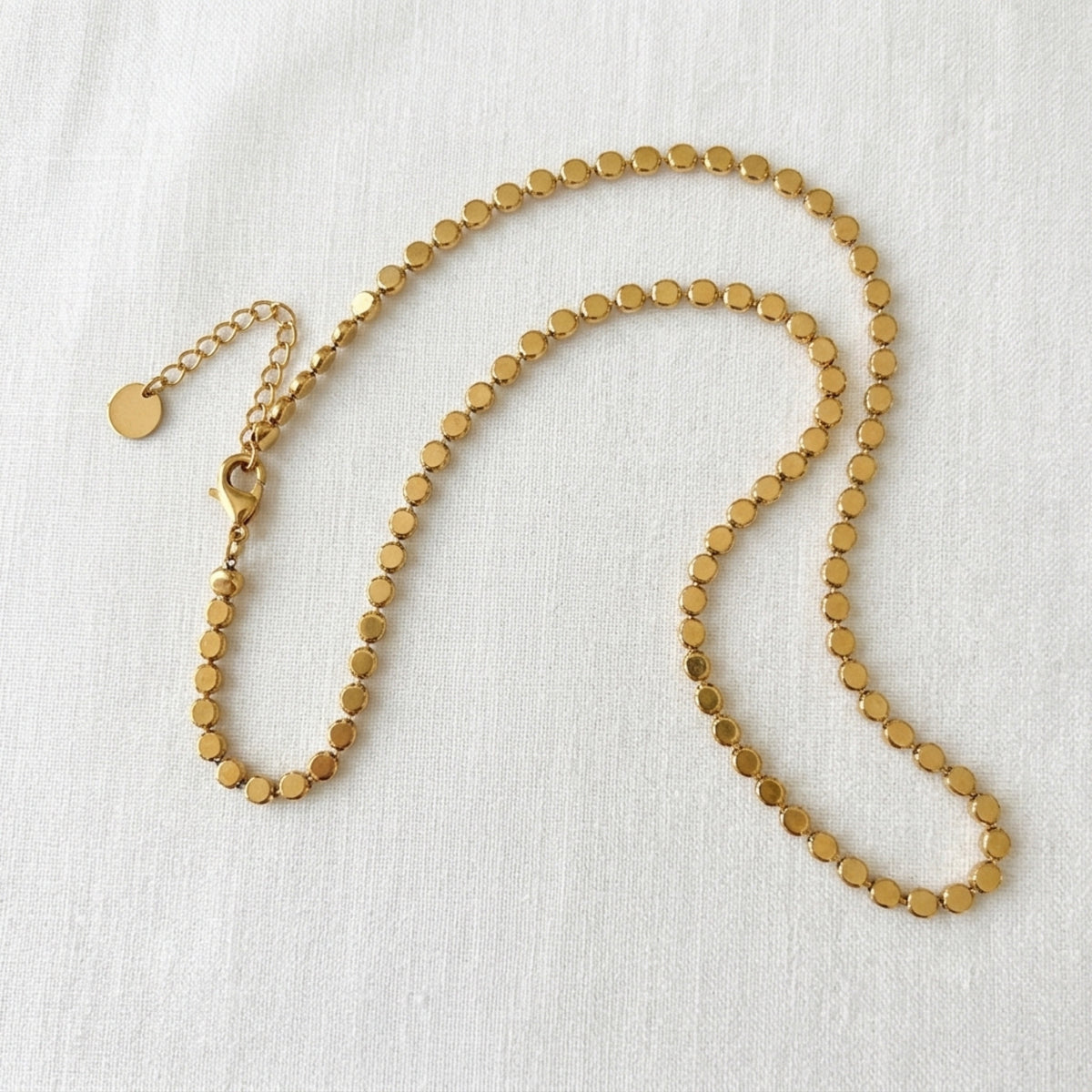 A gold-toned beaded chain necklace with flat circular beads and an adjustable lobster clasp with a small round tag, displayed on a textured white fabric background.  Golden Mirage Sequin Necklace - The Songbird Collection
