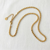 A gold-toned beaded chain necklace with flat circular beads and an adjustable lobster clasp with a small round tag, displayed on a textured white fabric background.  Golden Mirage Sequin Necklace - The Songbird Collection