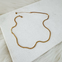 A delicate gold chain necklace with small, faceted cube-like beads, displayed on a textured white fabric surface.  Golden Mirage Sequin Necklace - The Songbird Collection