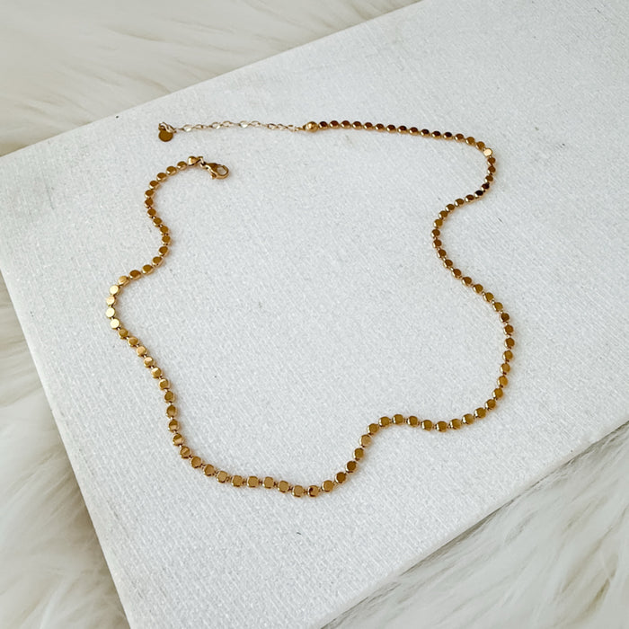 A delicate gold chain necklace with small, faceted cube-like beads, displayed on a textured white fabric surface.  Golden Mirage Sequin Necklace - The Songbird Collection