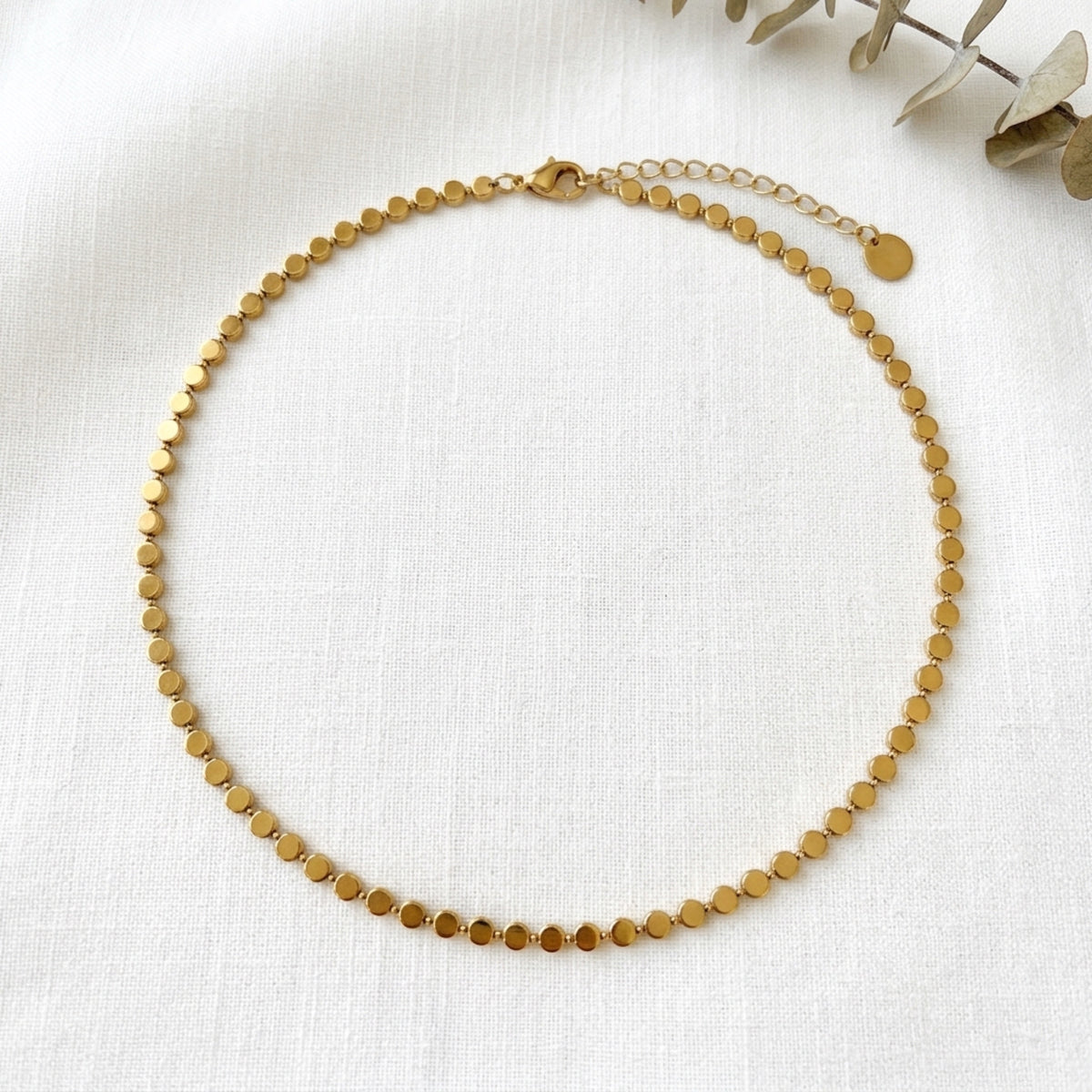 A delicate gold chain anklet with small, flat circular discs linked together, displayed on a textured white fabric background with dried eucalyptus leaves in the corner. Golden Mirage Sequin Necklace - The Songbird Collection