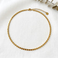 A delicate gold chain anklet with small, flat circular discs linked together, displayed on a textured white fabric background with dried eucalyptus leaves in the corner. Golden Mirage Sequin Necklace - The Songbird Collection