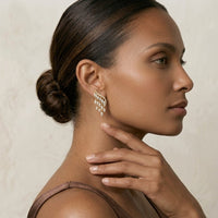 A close-up portrait of a woman wearing a delicate gold  Grace Chandelier Earrings - The Songbird Collection  set against a soft beige background.