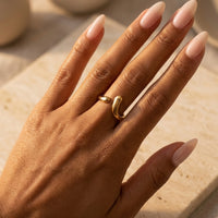 Hand wearing a gold ring with beige nail polish on a neutral background  Liquid Curve Ring - The Songbird Collection