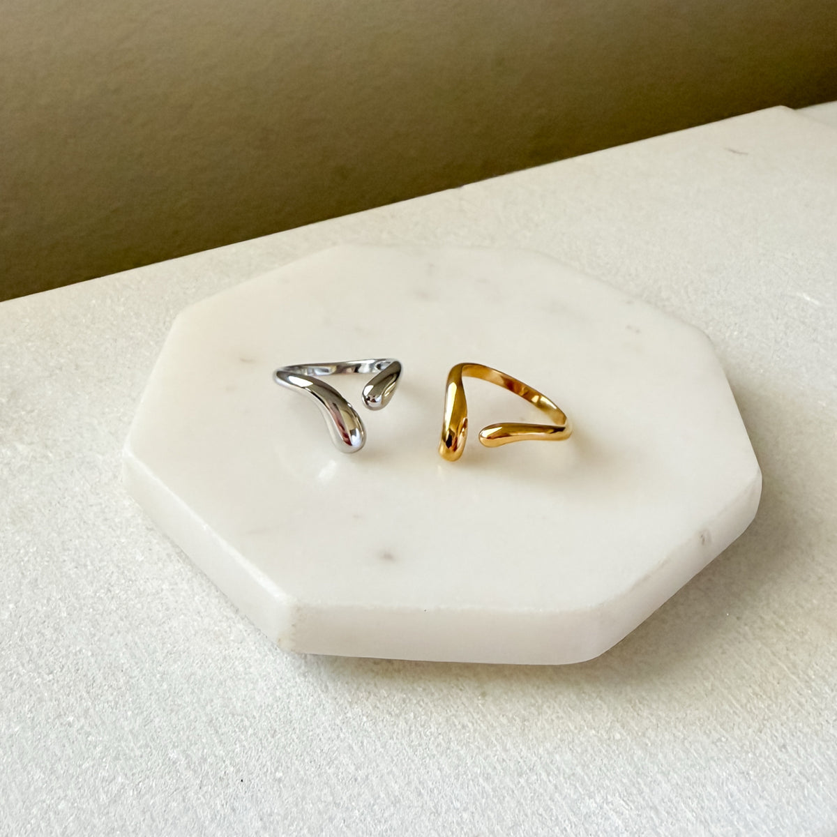 Two rings, one gold and one silver, on a white marble surface with a beige background.  Liquid Curve Ring - The Songbird Collection