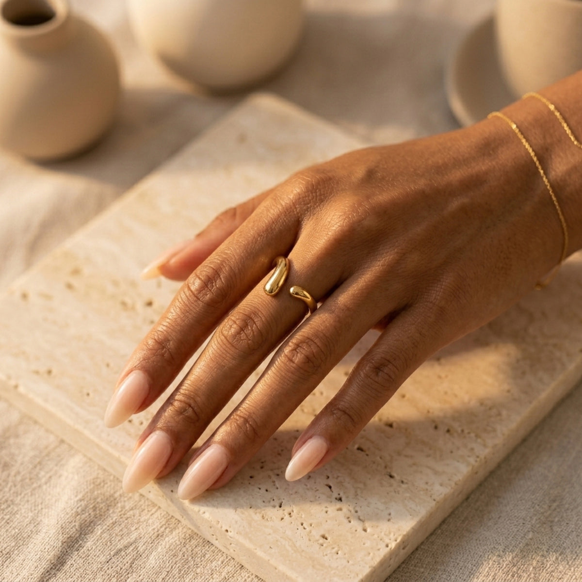 Hand wearing a gold ring on a textured surface with ceramic items in the background  Liquid Curve Ring - The Songbird Collection