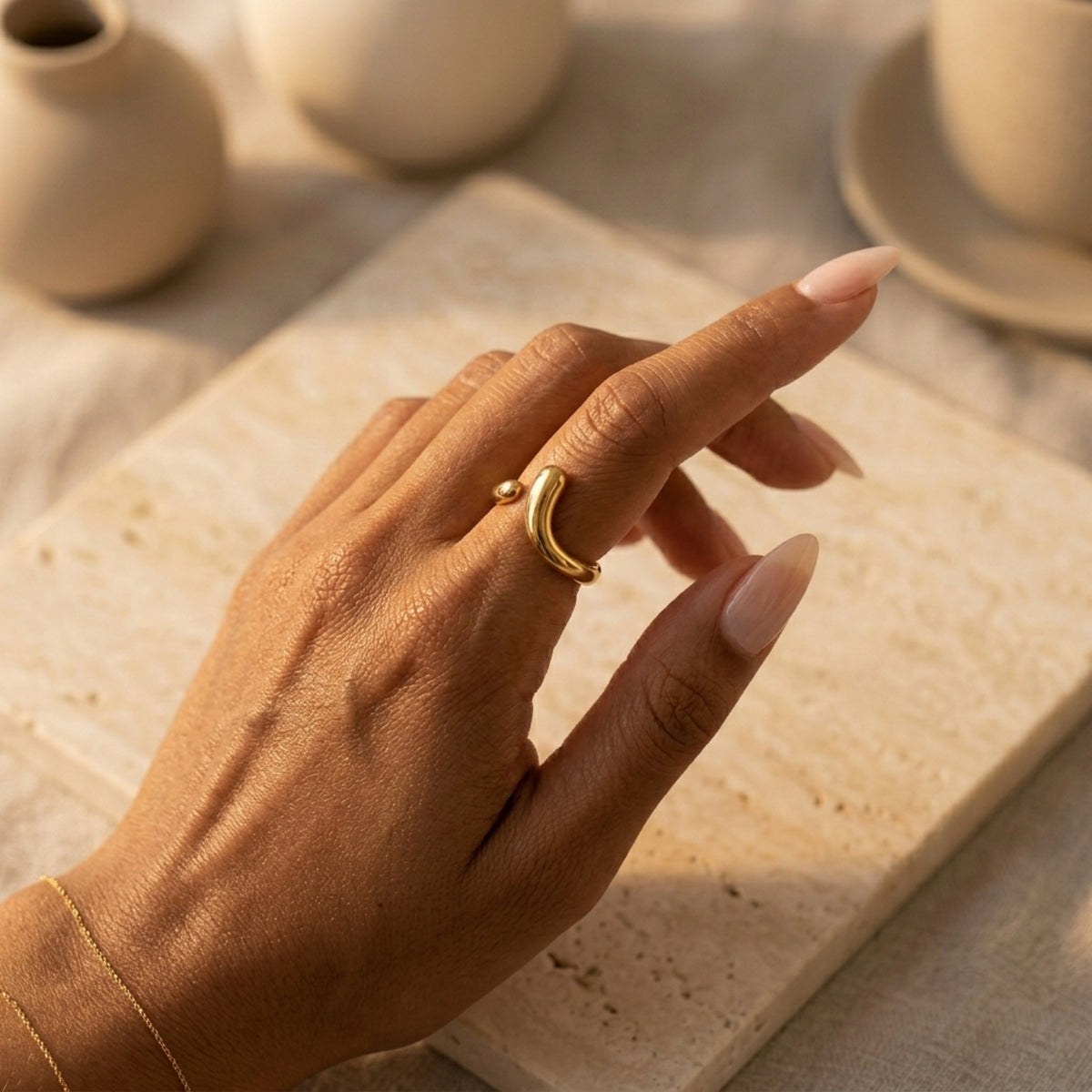 Hand wearing a gold ring on a textured surface with ceramic items in the  background Liquid Curve Ring - The Songbird Collection