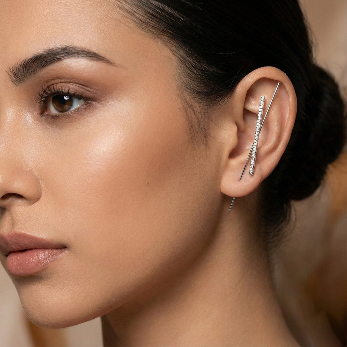Close-up of a woman's ear with a diamond earring against a neutral Woman wearing a silver ear cuff/ ear hook/ earring with a a bar of micropave setting of diamonds