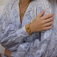 A person wears a light blue and white paisley blouse with lace details, accessorized with The Songbird Collection’s Icon Link Necklace and matching bracelet, both featuring chunky gold links and oversized clasps.