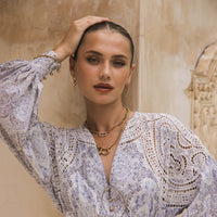 A woman with slicked-back hair poses with one arm raised, wearing a long-sleeved patterned blouse and The Songbird Collections Icon Link Necklace, standing against a textured beige wall.