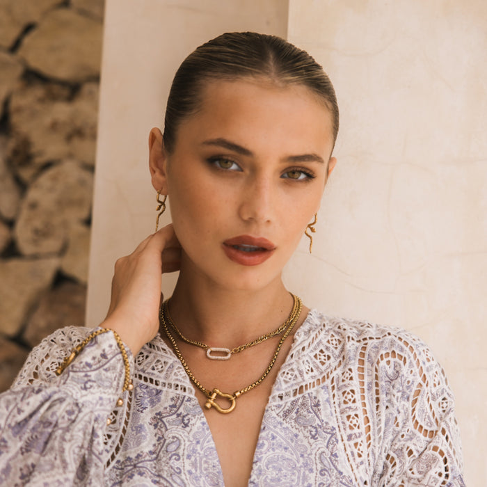 A woman with slicked-back hair models The Songbird Collection’s Icon Link Necklace, styled with a white patterned blouse, posing with one hand near her neck against a light stone wall and stacked rocks in the background.