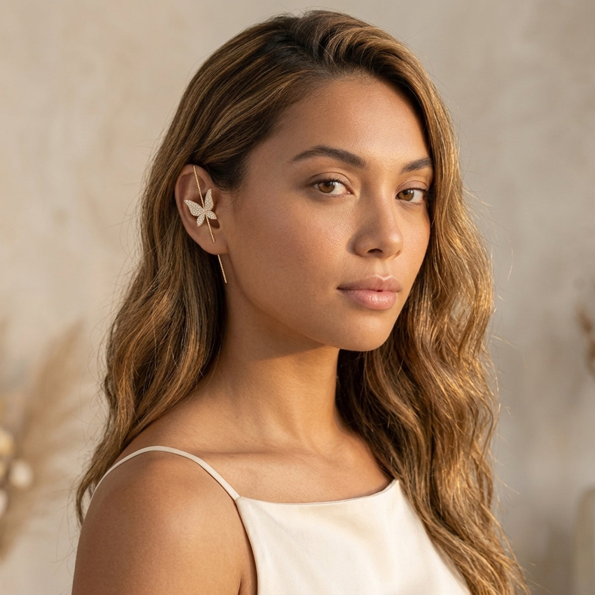 A woman with long wavy brown hair wearing a delicate gold ear cuff with a butterfly design, paired with a white camisole top against a soft beige background.